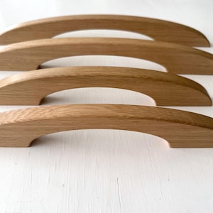 Puede incluir: Cuatro tiradores de madera curvados, probablemente de roble claro. Los tiradores tienen un acabado liso y pulido, con un diseño arqueado. Están dispuestos en fila, mostrando su forma y la veta natural de la madera.