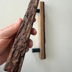 May include: Two wooden door pulls with a natural bark texture. One is held in a hand, showcasing the detailed, rough surface. The other is mounted, revealing a dark green cylindrical hardware. The wood tones range from light to dark brown.
