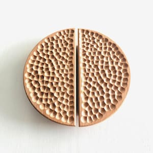 May include: A pair of wooden cabinet pulls, each in a half-circle shape. The surface of each pull is intricately carved with a pattern of small, irregular shapes. The wood is light brown, and the pulls are set against a white background.