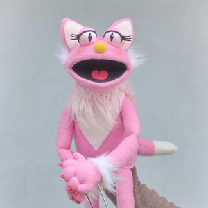 May include: A pink cat puppet with white fur accents and red claws. The puppet has large eyes with long eyelashes, a yellow nose, and a wide open mouth. The puppet is being held by a person.
