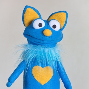 May include: A blue puppet with yellow ears, nose, and a heart on its chest. The puppet has a fluffy blue collar and large white eyes with black pupils. The puppet's arms are blue, and the bottom portion of the puppet is held by a person wearing a black and white striped sleeve.