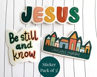 May include: A sticker pack of three designs. The first sticker features the word "JESUS" in colorful letters. The second sticker features the phrase "Be still and know" in green and yellow lettering. The third sticker features a row of colorful houses with the text "Love your neighbor" and a Bible verse reference.