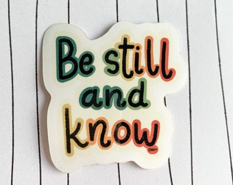 May include: A sticker with the text "Be still and know" in a rainbow color scheme. The sticker has a white background and a slightly rounded shape.