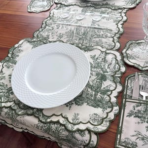 May include: A dining table setting with a white plate, a wine glass, and a placemat with a green and white toile pattern. The placemat, coasters, and cutlery holder feature a scalloped edge.