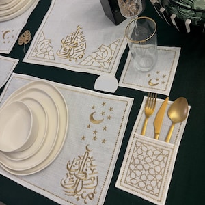 Op de afbeelding: Witte linnen placemats met gouden borduurwerk van islamitische kalligrafie en sterpatronen. De placemats liggen op een donkergroene ondergrond met witte borden, een glas en gouden bestek.