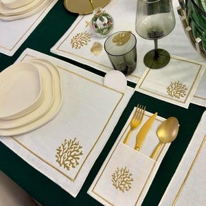 Può includere: Tovagliette di lino bianco con disegni di corallo ricamati in oro. Le tovagliette sono disposte su una tovaglia verde scuro. Ci sono anche portaposate dorati con un disegno di corallo ricamato in oro.