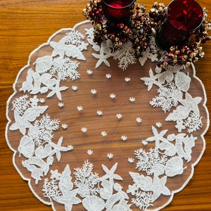 Może przedstawiać: Biały bieżnik stołowy z koronki z falowanym brzegiem i haftowanym wzorem kwiatowym. Bieżnik leży na drewnianym stole z dwoma czerwonymi szklanymi świecznikami.