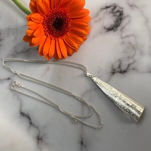 May include: A silver-toned necklace with a textured, triangular pendant. The chain is delicate and the pendant has a brushed metal appearance. An orange gerbera daisy rests in the background, adding a pop of color.