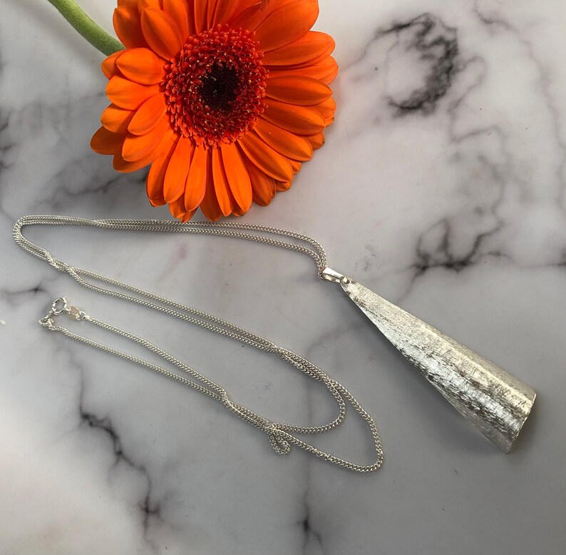 May include: A silver-toned necklace with a textured, triangular pendant. The chain is delicate and the pendant has a brushed metal appearance. An orange gerbera daisy rests in the background, adding a pop of color.