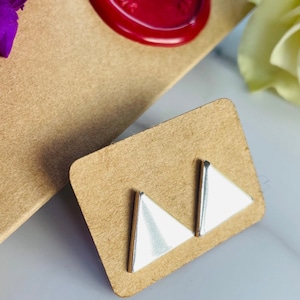 May include: Silver-tone triangle stud earrings with white enamel, presented on a small brown card. The earrings have a minimalist design. The card is on a marble surface, with a red wax seal and purple flower visible.