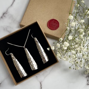 May include: A silver jewelry set featuring a necklace and matching earrings with a hammered texture. The set is displayed in a black-lined box with a brown exterior, alongside a sprig of white flowers. A red wax seal is visible on the box.