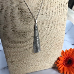 May include: A silver-toned pendant necklace with a textured, cone-shaped pendant. The pendant hangs from a delicate chain. An orange gerbera daisy is in the background.