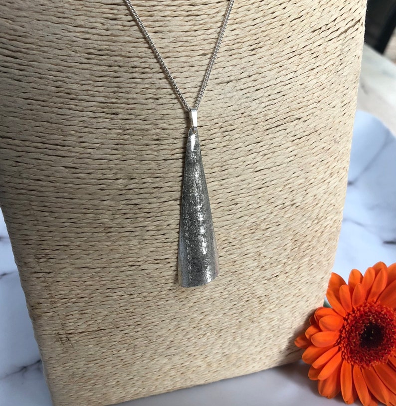 May include: A silver-toned pendant necklace with a textured, cone-shaped pendant. The pendant hangs from a delicate chain. An orange gerbera daisy is in the background.