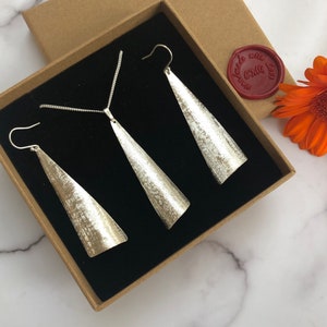 May include: A silver jewelry set, including a necklace and a pair of earrings, presented in a brown box with a black interior. The earrings and pendant have a textured, cone-like design. A red wax seal with text is visible.