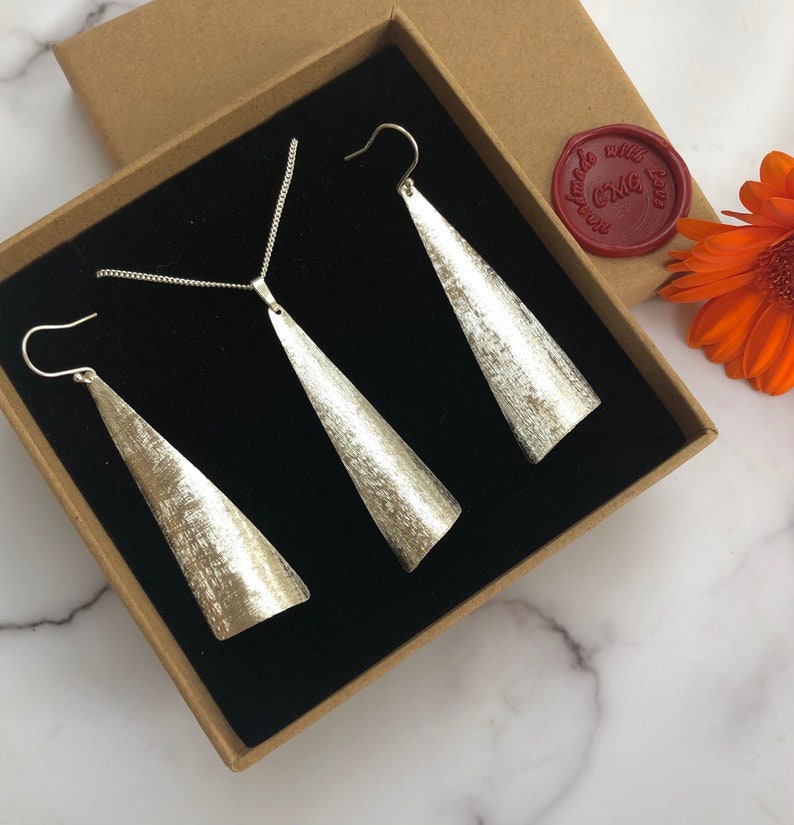 May include: A silver jewelry set, including a necklace and a pair of earrings, presented in a brown box with a black interior. The earrings and pendant have a textured, cone-like design. A red wax seal with text is visible.