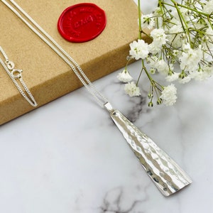 May include: A silver pendant necklace with a hammered texture. The pendant is a long, asymmetrical shape. The necklace is displayed on a marble surface with white flowers and a red wax seal in the background.