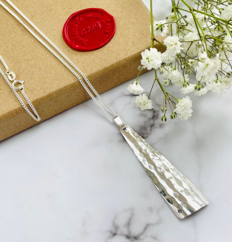 May include: A silver pendant necklace with a hammered texture. The pendant is a long, asymmetrical shape. The necklace is displayed on a marble surface with white flowers and a red wax seal in the background.