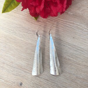 May include: A pair of silver-toned, cone-shaped earrings with a textured surface. The earrings hang from silver hooks. A large, red flower with green leaves is in the background, resting on a light wood surface.