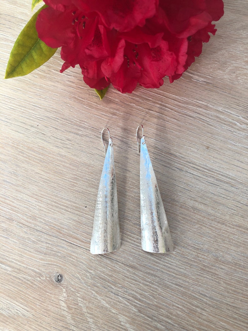 May include: A pair of silver-toned, cone-shaped earrings with a textured surface. The earrings hang from silver hooks. A large, red flower with green leaves is in the background, resting on a light wood surface.