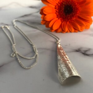 May include: A silver necklace with a textured, cone-shaped pendant. The necklace chain is delicate and hangs on a marble surface, with an orange gerbera daisy in the background.
