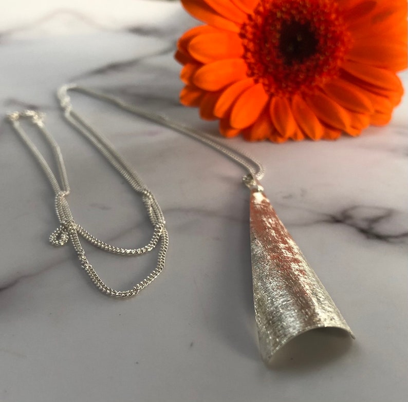 May include: A silver necklace with a textured, cone-shaped pendant. The necklace chain is delicate and hangs on a marble surface, with an orange gerbera daisy in the background.