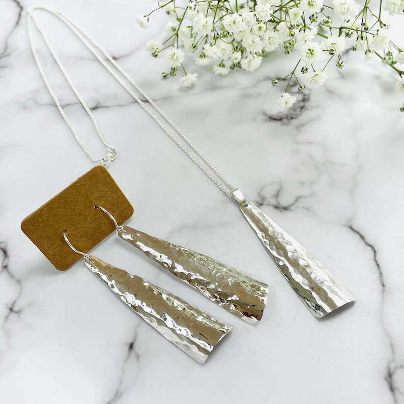 May include: A silver jewelry set featuring a hammered texture. The set includes a long pendant necklace and a pair of dangle earrings. The jewelry is displayed on a white marble surface with white flowers in the background.
