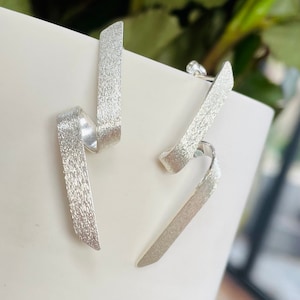 May include: A pair of silver earrings with a textured finish. The earrings have a unique, abstract design, with a ribbon-like shape that curves and twists. The earrings are displayed on a white surface, with a blurred green background.