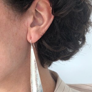 May include: Silver-toned, textured earrings with a long, tapered design. The earrings are suspended from a simple hook and are worn by a person with dark hair. The earrings have a brushed metal appearance.