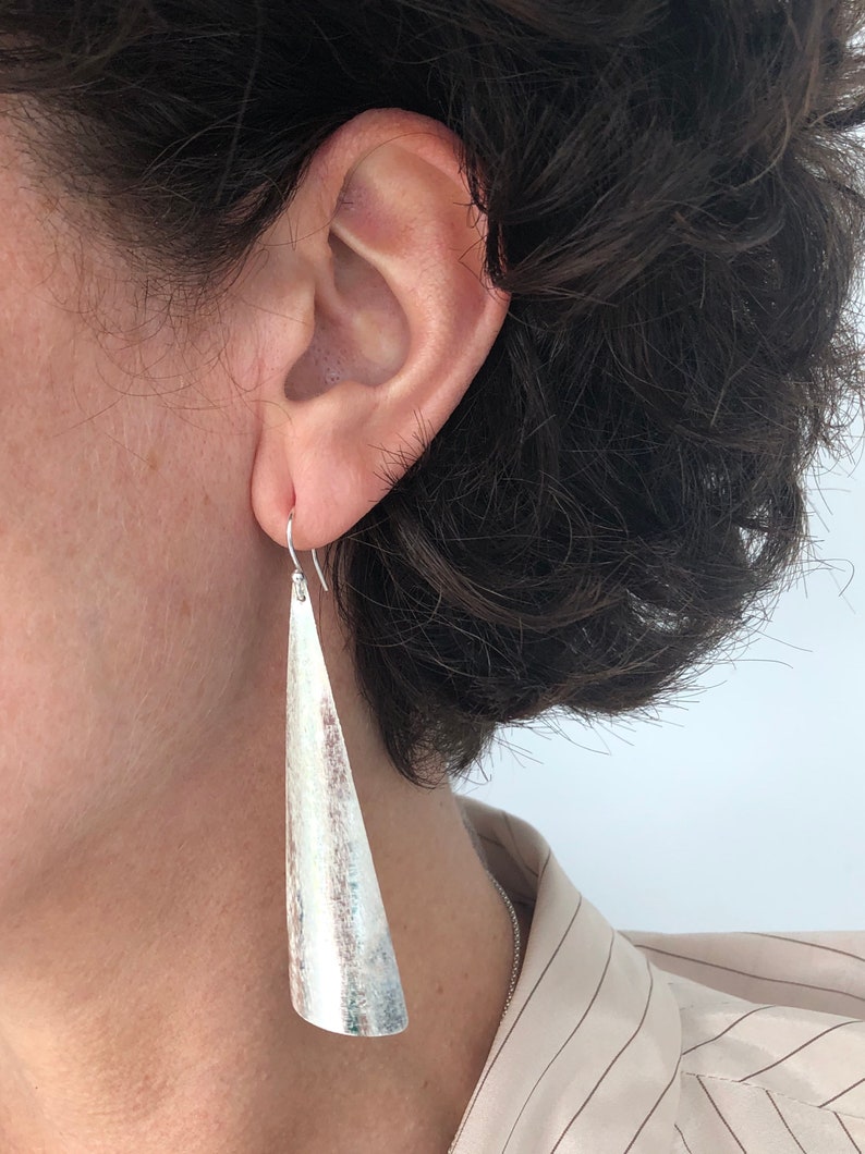 May include: Silver-toned, textured earrings with a long, tapered design. The earrings are suspended from a simple hook and are worn by a person with dark hair. The earrings have a brushed metal appearance.