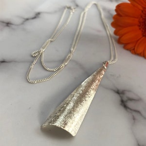 May include: A silver-toned necklace with a textured, cone-shaped pendant. The pendant hangs from a delicate chain, and the necklace is displayed on a marble surface. An orange flower is visible in the background.