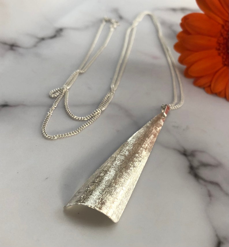 May include: A silver-toned necklace with a textured, cone-shaped pendant. The pendant hangs from a delicate chain, and the necklace is displayed on a marble surface. An orange flower is visible in the background.
