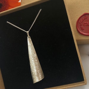 May include: A silver-toned pendant necklace with a textured, elongated cone-shaped pendant. The pendant hangs from a delicate chain and is displayed in a black velvet-lined box. The necklace is presented in a cardboard box with a red wax seal.