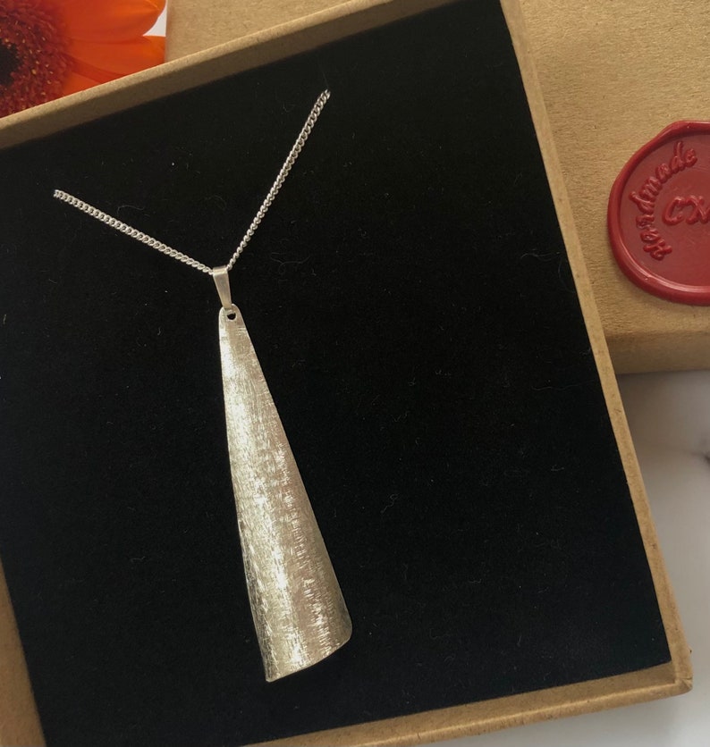 May include: A silver-toned pendant necklace with a textured, elongated cone-shaped pendant. The pendant hangs from a delicate chain and is displayed in a black velvet-lined box. The necklace is presented in a cardboard box with a red wax seal.