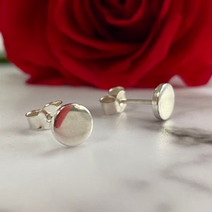 May include: Two small, round, silver stud earrings. The earrings have a simple, minimalist design with a flat, circular face. The earrings are displayed on a white surface with a blurred red rose in the background.