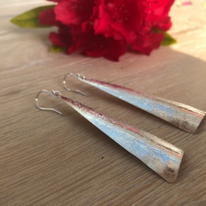 May include: A pair of silver-toned, long, triangular earrings with a brushed metal finish. The earrings have a hook closure and are displayed on a wooden surface, with a red flower in the background.