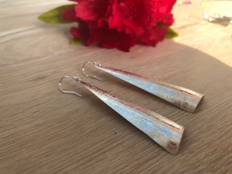 May include: A pair of silver-toned, long, triangular earrings with a brushed metal finish. The earrings have a hook closure and are displayed on a wooden surface, with a red flower in the background.