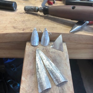 May include: Silver-toned, handcrafted earrings and earring components on a wooden workbench. The earrings are cone-shaped with textured surfaces. Additional metal pieces and tools are visible in the background, suggesting a jewelry-making setting.