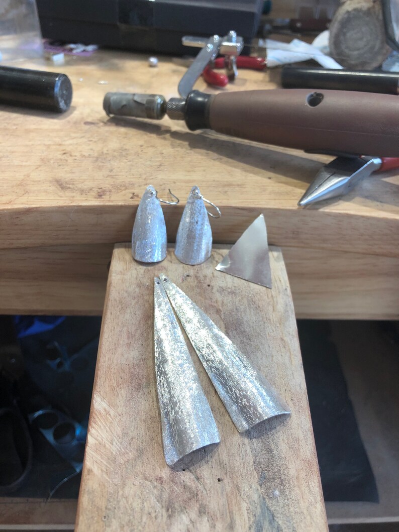 May include: Silver-toned, handcrafted earrings and earring components on a wooden workbench. The earrings are cone-shaped with textured surfaces. Additional metal pieces and tools are visible in the background, suggesting a jewelry-making setting.
