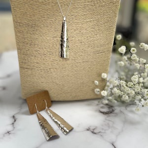 May include: A silver pendant necklace and matching earrings. The pendant is a long, textured, trapezoid shape hanging from a silver chain. The earrings are a similar trapezoid shape with a hammered texture. The jewelry is displayed on a neutral-toned bust.