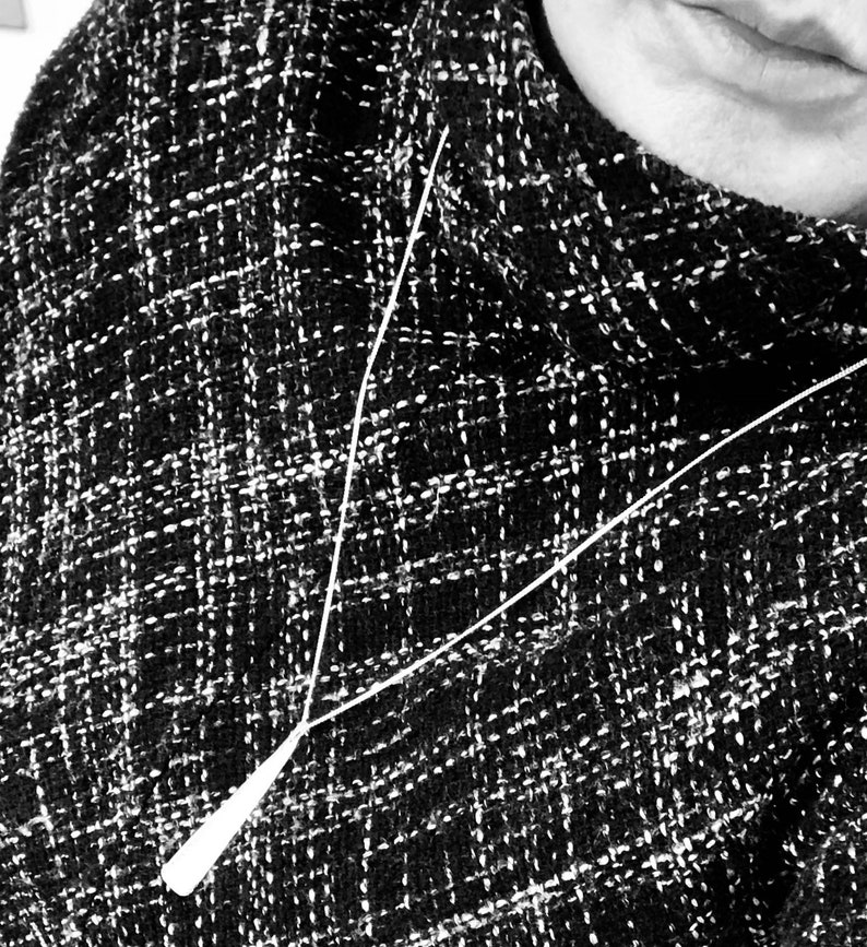 May include: Black and white tweed fabric with a white pendant necklace. The necklace has a long, thin chain and a teardrop-shaped pendant. The tweed fabric has a woven pattern.