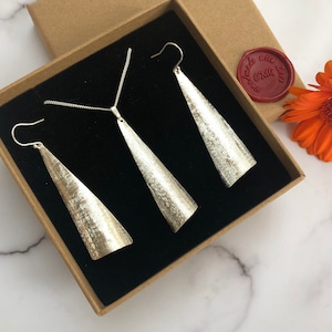 May include: A silver jewelry set, including a necklace and two earrings, presented in a brown gift box with a black interior. The jewelry has a textured, brushed metal finish. A red wax seal and an orange flower are also visible.