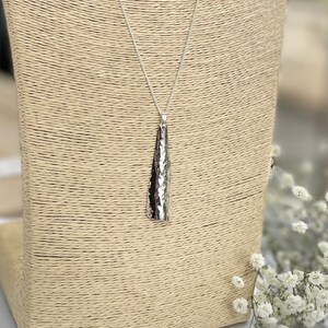May include: A silver pendant necklace with a textured, cone-shaped pendant. The pendant hangs from a delicate chain. The necklace is displayed on a beige jewelry stand, with white flowers in the background.