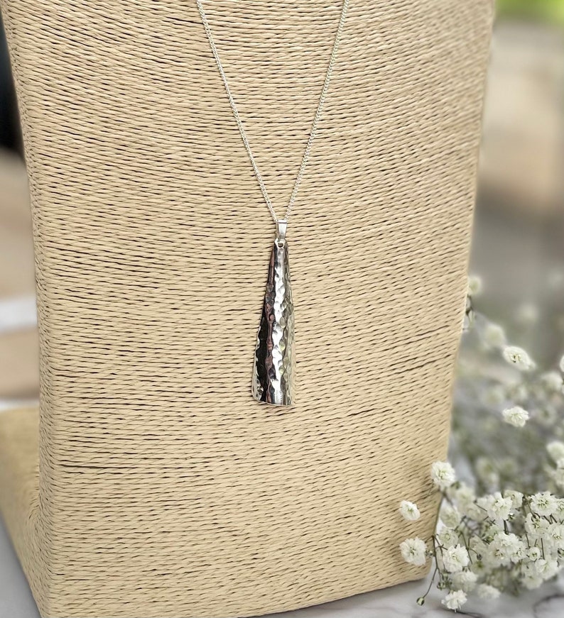 May include: A silver pendant necklace with a textured, cone-shaped pendant. The pendant hangs from a delicate chain. The necklace is displayed on a beige jewelry stand, with white flowers in the background.