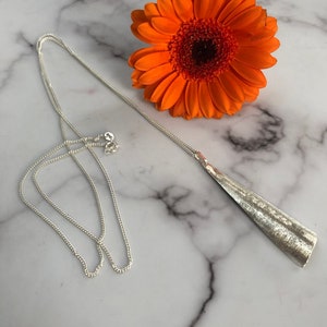 May include: A silver necklace with a long, textured pendant in a triangular shape. The necklace is displayed on a marble surface next to a bright orange gerbera daisy.