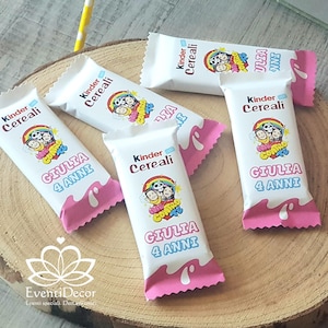 Può includere: Diverse barrette di cioccolato Kinder Cereali bianche con dettagli rosa, disposte su una superficie di legno. Le barrette presentano una grafica a cartoni animati e il nome "GIULIA" con "4 ANNI" sotto.