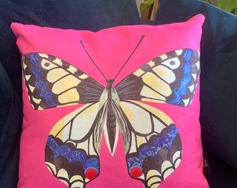 May include: A pink pillow with a large butterfly design. The butterfly has black, white, blue, and red accents. The pillow is made of a soft, plush fabric.
