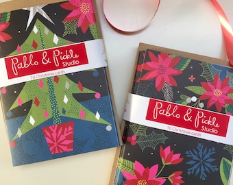 May include: Two packs of Christmas cards featuring festive designs. The first pack has a green Christmas tree with a pink pot on a dark blue background. The second pack has red poinsettias and snowflakes on a black background. Both packs have a brown paper envelope and a white label with the text "Pablo & Pickle Studio" and "10 Christmas cards".