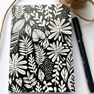 May include: Black and white notebook with a floral pattern. The cover features a repeating design of leaves and flowers in a white silhouette against a black background.
