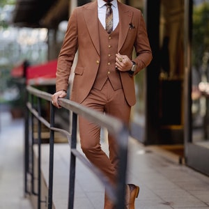 May include: A man wearing a brown three-piece suit with a white shirt and a patterned tie. He is standing on a sidewalk and holding onto a railing.