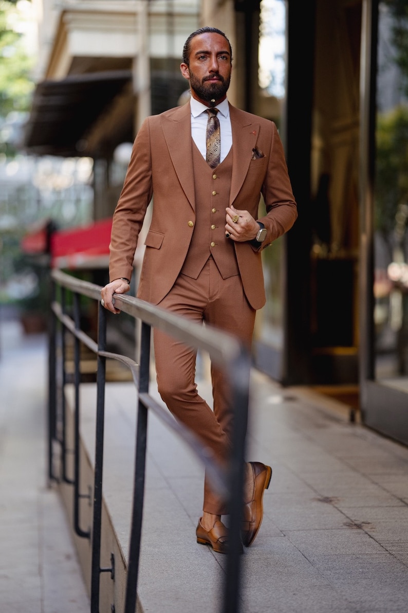 May include: A man wearing a brown three-piece suit with a white shirt and a patterned tie. He is standing on a sidewalk and holding onto a railing.